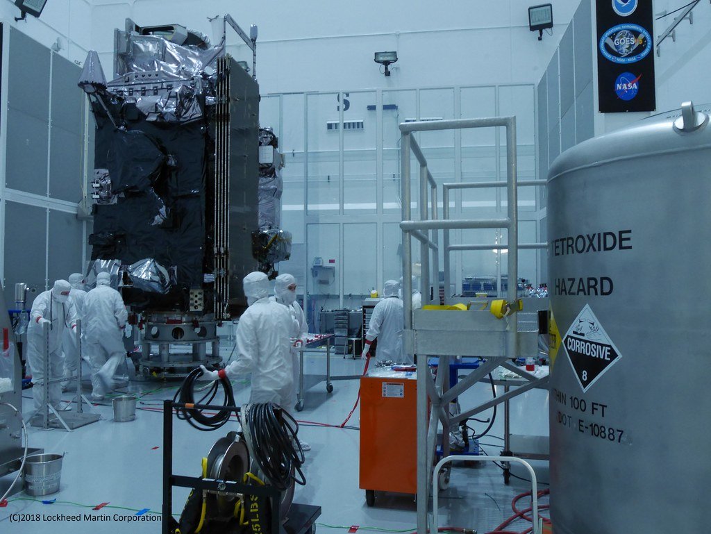 A worker in a hard hat moves a 55-gallon steel drum onto grated secondary containment flooring. A 'NON RCRA WASTE' sign is visible on the wall, indicating the area is segregated by waste classification.