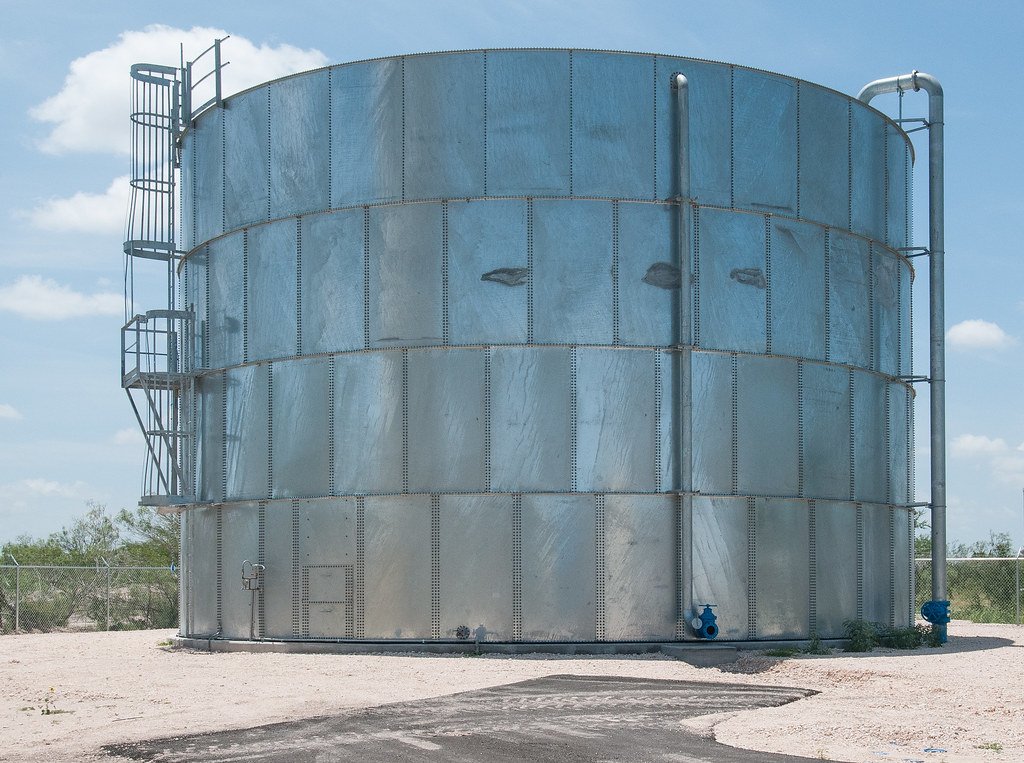 Three white aboveground bulk fuel storage tanks surrounded by an earthen and gravel secondary containment berm, with transfer piping crossing the dike. The containment area is graded to capture any spill from the tanks.