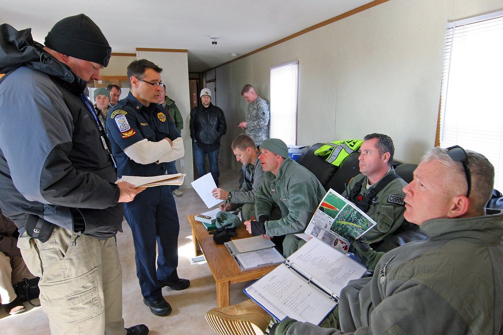 Two firefighters in uniform consult a spill response checklist together, focused on the document, with response equipment visible in the foreground.